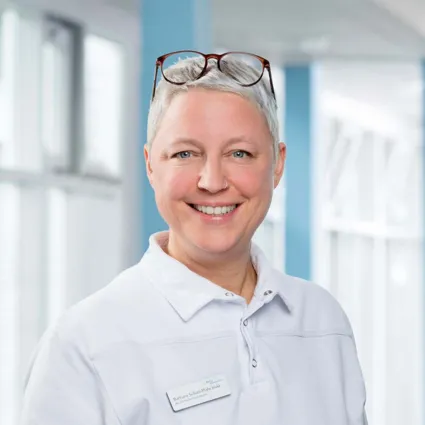 Lächelnde Frau mit kurzen grauen Haaren, trägt ein weißes Oberteil und eine Namensschilder, im Hintergrund helle, moderne Umgebung mit Fenstern und blauen Wänden.