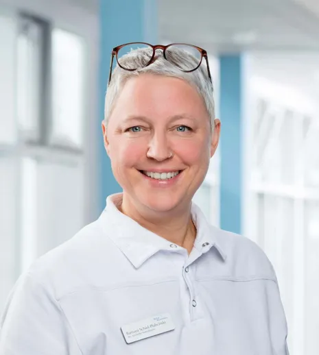 Lächelnde Frau mit kurzen grauen Haaren, trägt ein weißes Oberteil und eine Namensschilder, im Hintergrund helle, moderne Umgebung mit Fenstern und blauen Wänden.