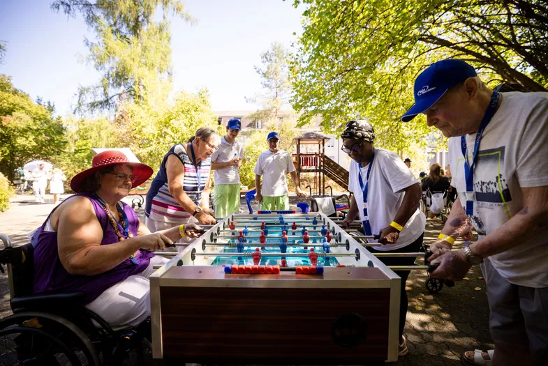 Menschen spielen im Freien Tischfußball; eine Frau im Rollstuhl und mehrere Mitspieler stehen an einem Kicker unter Bäumen.