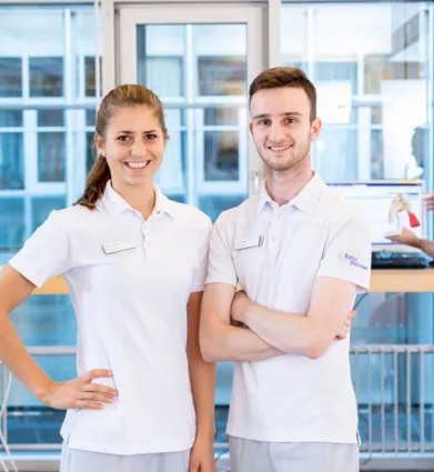 Zwei Mitarbeiter in weißen Poloshirts stehen in einem modernen Empfangsbereich. Die Frau lächelt und hat eine Hand an der Hüfte, der Mann steht mit verschränkten Armen. Im Hintergrund ist ein Computer mit einem Bildschirm sichtbar, auf dem eine medizin...