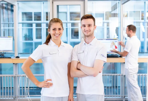 Zwei Mitarbeiter in weißen Poloshirts stehen in einem modernen Empfangsbereich. Die Frau lächelt und hat eine Hand an der Hüfte, der Mann steht mit verschränkten Armen. Im Hintergrund ist ein Computer mit einem Bildschirm sichtbar, auf dem eine medizin...