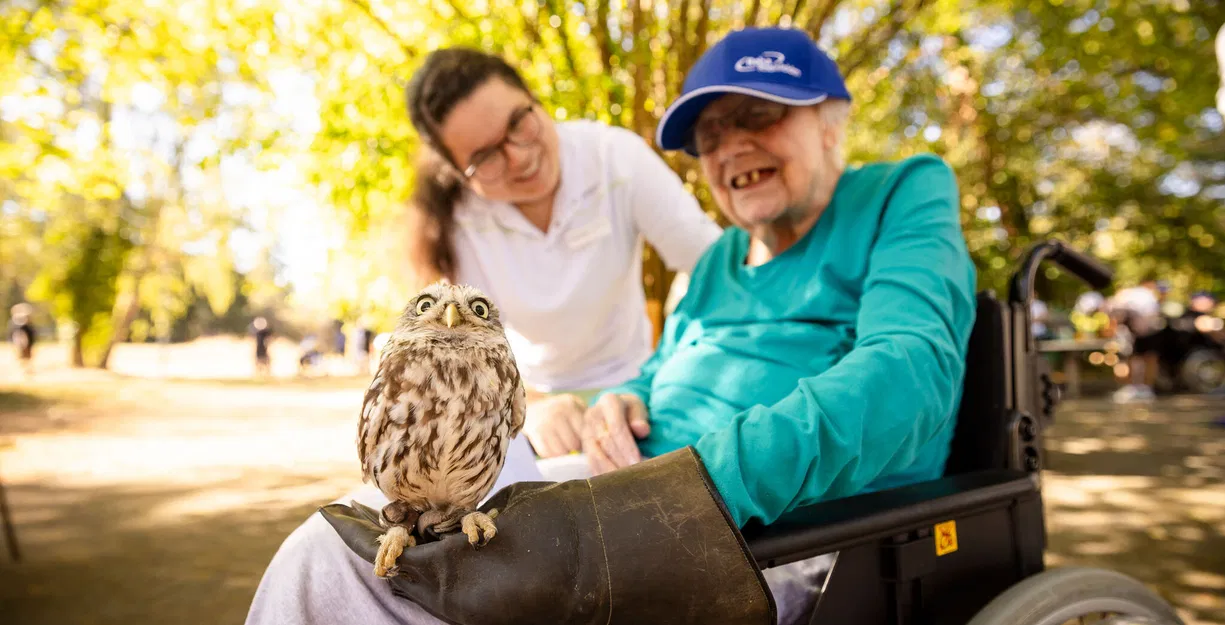 Seniorin im Rollstuhl mit Eule auf dem Schoß, begleitet von einer Frau in einem sonnigen Park.