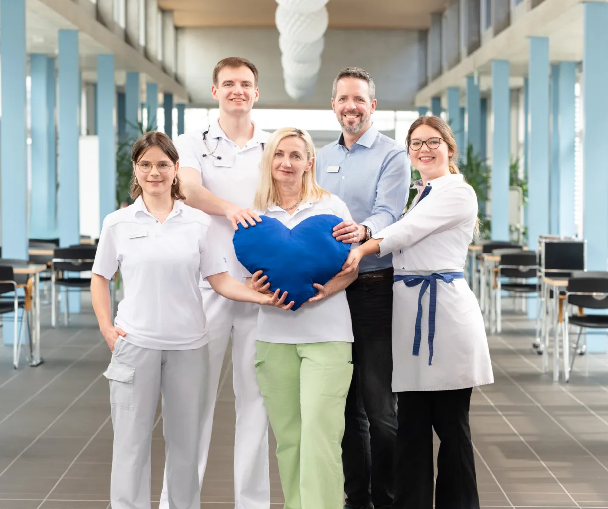 Fünf Personen stehen in einem modernen Raum mit großen Fenstern. Sie tragen unterschiedliche Arbeitskleidung, darunter medizinische Uniformen und eine weiße Schürze. In der Mitte halten sie ein großes, blaues Herz. Der Raum hat einen hellen Boden und i...