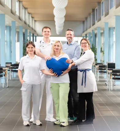 Fünf Personen stehen in einem modernen Raum mit großen Fenstern. Sie tragen unterschiedliche Arbeitskleidung, darunter medizinische Uniformen und eine weiße Schürze. In der Mitte halten sie ein großes, blaues Herz. Der Raum hat einen hellen Boden und i...