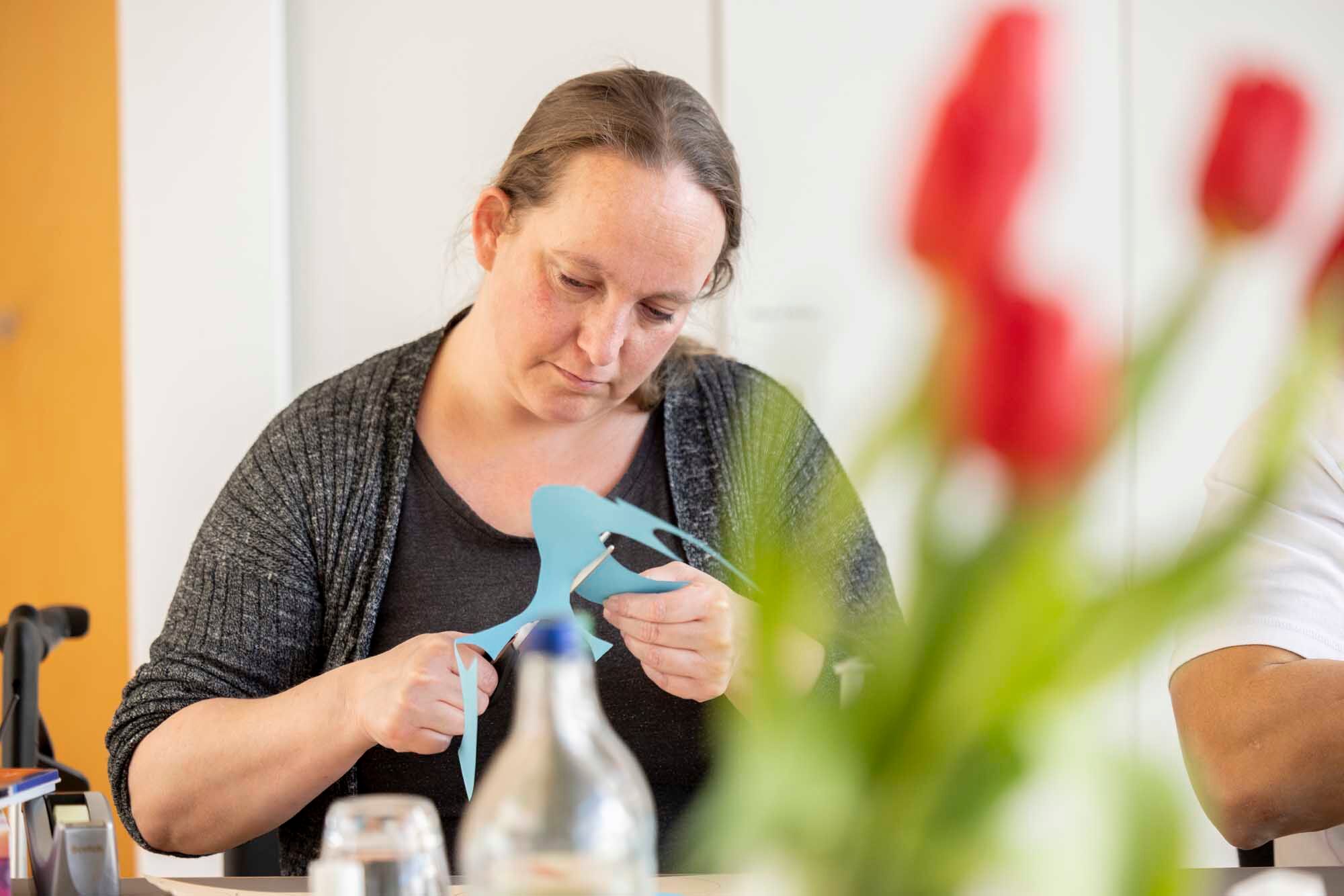 Frau mit langen Haaren schneidet mit einer Schere eine blaue Papierfigur aus. Im Vordergrund sind verschwommen rote Blumen und ein Glas Wasser sichtbar.