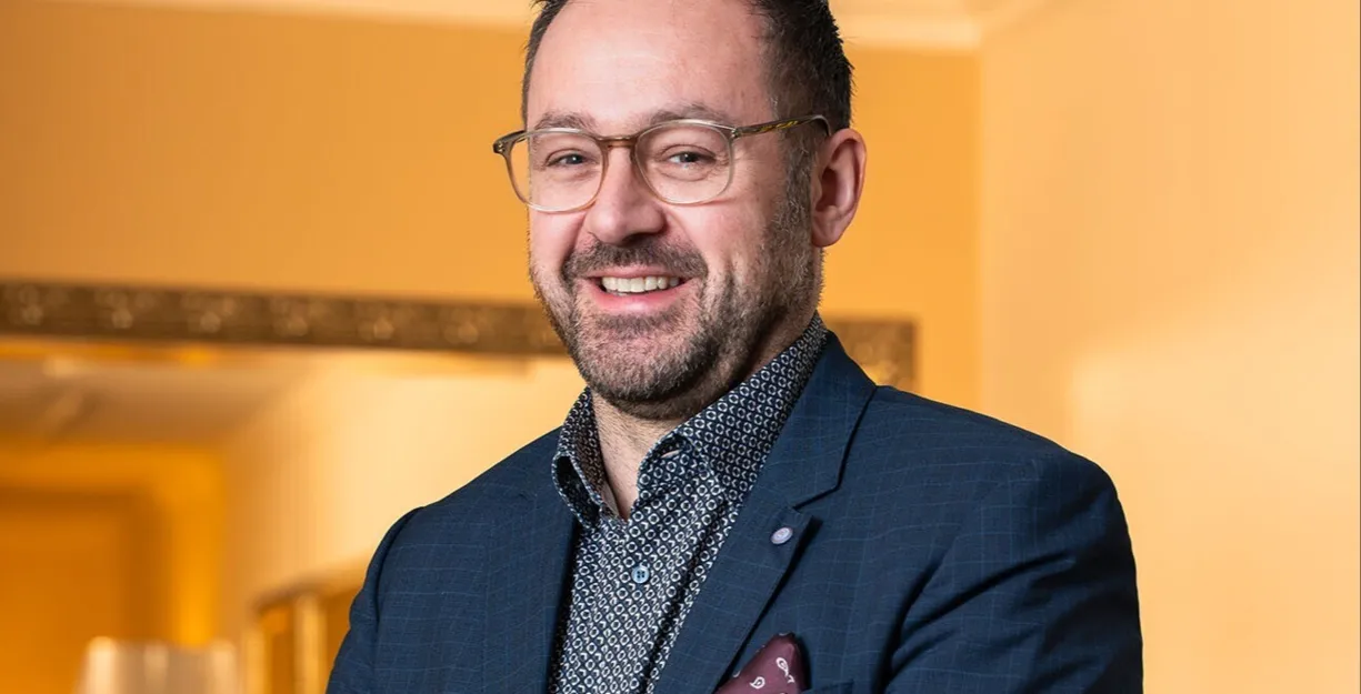 Mann mit Brille und Bart, gekleidet in einem blauen Anzug mit gemusterter Hemd, lächelt und steht vor einer hellen Wand mit einem Spiegel im Hintergrund. Ein Namensschild ist an seiner Jacke befestigt.