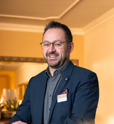 Mann mit Brille und Bart, gekleidet in einem blauen Anzug mit gemusterter Hemd, lächelt und steht vor einer hellen Wand mit einem Spiegel im Hintergrund. Ein Namensschild ist an seiner Jacke befestigt.