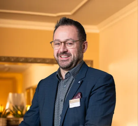 Mann mit Brille und Bart, gekleidet in einem blauen Anzug mit gemusterter Hemd, lächelt und steht vor einer hellen Wand mit einem Spiegel im Hintergrund. Ein Namensschild ist an seiner Jacke befestigt.