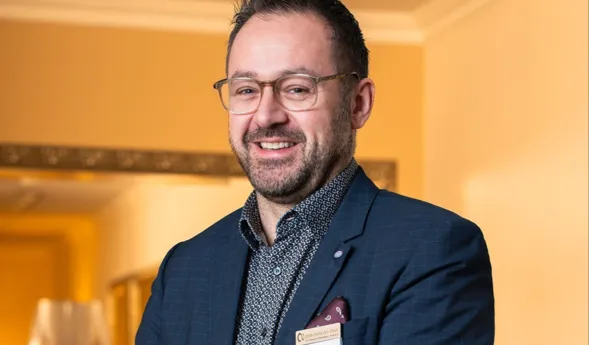 Mann mit Brille und Bart, gekleidet in einem blauen Anzug mit gemusterter Hemd, lächelt und steht vor einer hellen Wand mit einem Spiegel im Hintergrund. Ein Namensschild ist an seiner Jacke befestigt.