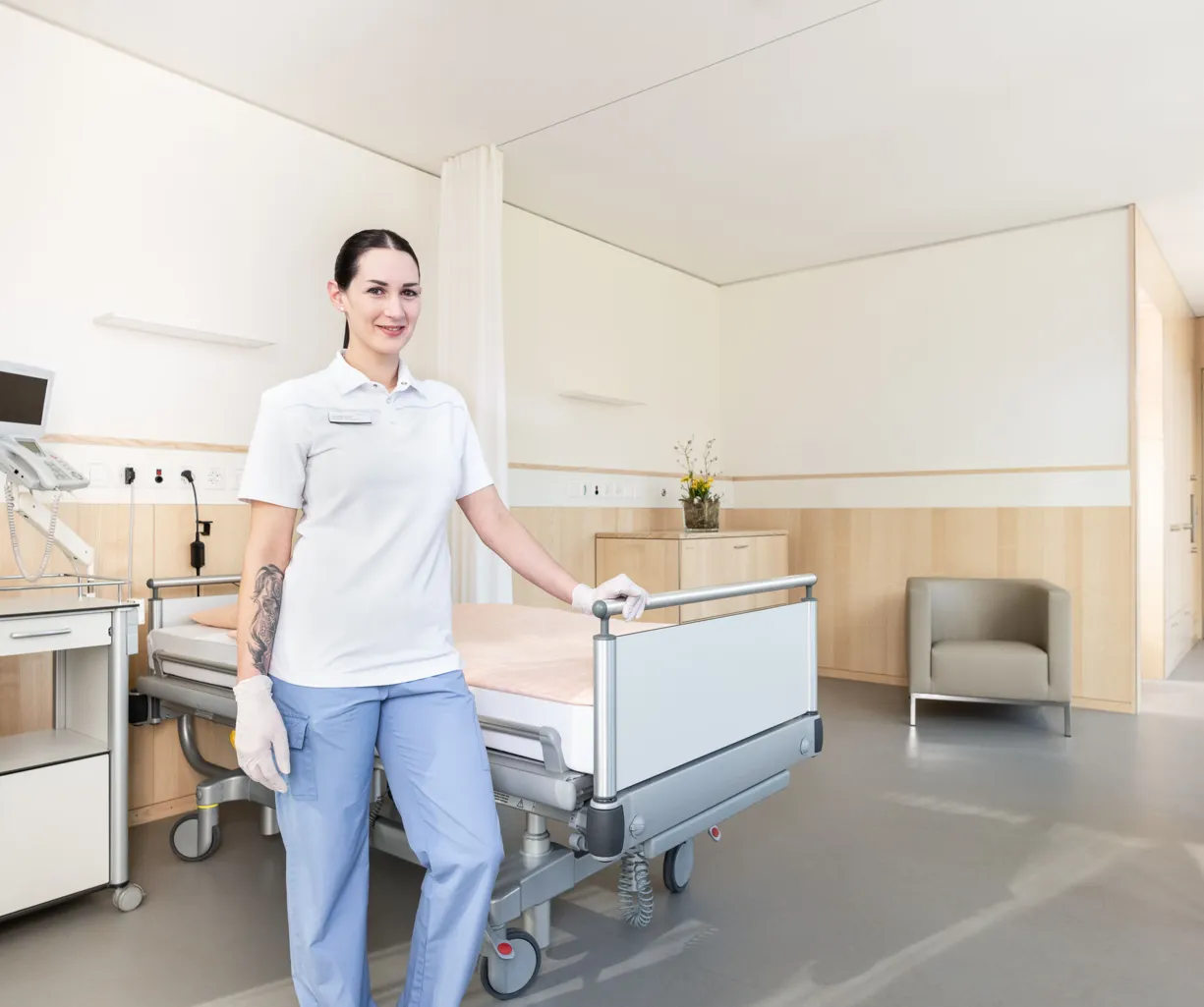 Pflegekraft in einem modernen Krankenhauszimmer, ausgestattet mit einem Bett, einem Nachttisch, einem Monitor und einem Sitzbereich. Helle Farben und Holzverkleidung schaffen eine freundliche Atmosphäre.