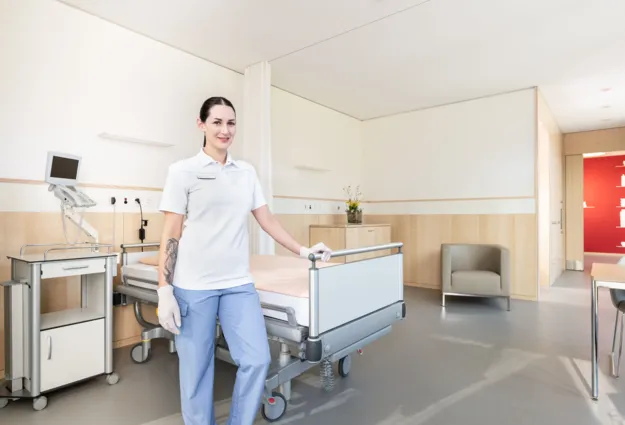 Pflegekraft in einem modernen Krankenhauszimmer, ausgestattet mit einem Bett, einem Nachttisch, einem Monitor und einem Sitzbereich. Helle Farben und Holzverkleidung schaffen eine freundliche Atmosphäre.