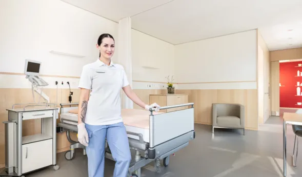 Pflegekraft in einem modernen Krankenhauszimmer, ausgestattet mit einem Bett, einem Nachttisch, einem Monitor und einem Sitzbereich. Helle Farben und Holzverkleidung schaffen eine freundliche Atmosphäre.