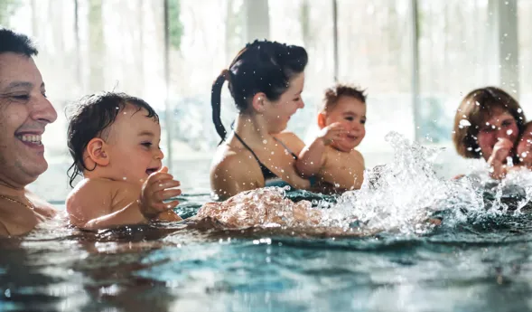 Vater und Mutter mit drei kleinen Kindern im Schwimmbecken, Planschen und Lachen, Wasser spritzt, warmes Licht durch große Fenster im Hintergrund.
