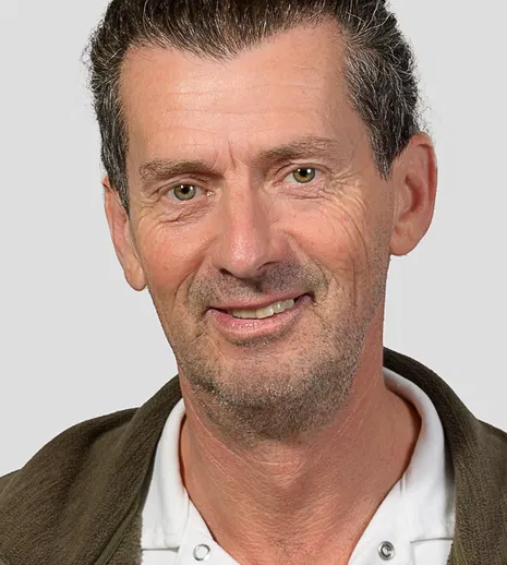 Porträt eines lächelnden, älteren Mannes mit dunklem, grau meliertem Haar, Bartstoppeln und grünen Augen vor grauem Hintergrund.
