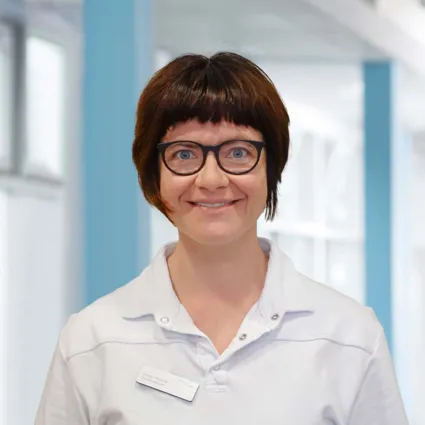 Frau mit kurzen, braunen Haaren und Brille, trägt ein weißes Oberteil. Lächelt freundlich vor einem hellen, modernen Hintergrund.