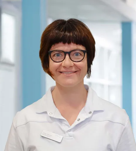 Frau mit kurzen, braunen Haaren und Brille, trägt ein weißes Oberteil. Lächelt freundlich vor einem hellen, modernen Hintergrund.