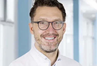 Lächelnder Mann mit Brille, Bart und weißem Kittel, vor einem hellen, modernen Hintergrund.