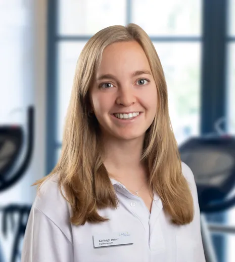 Lächelnde Frau im weißen Kittel vor Fitnessgeräten in einem hellen Raum mit großen Fenstern, im Hintergrund Crosstrainer und Fahrräder.
