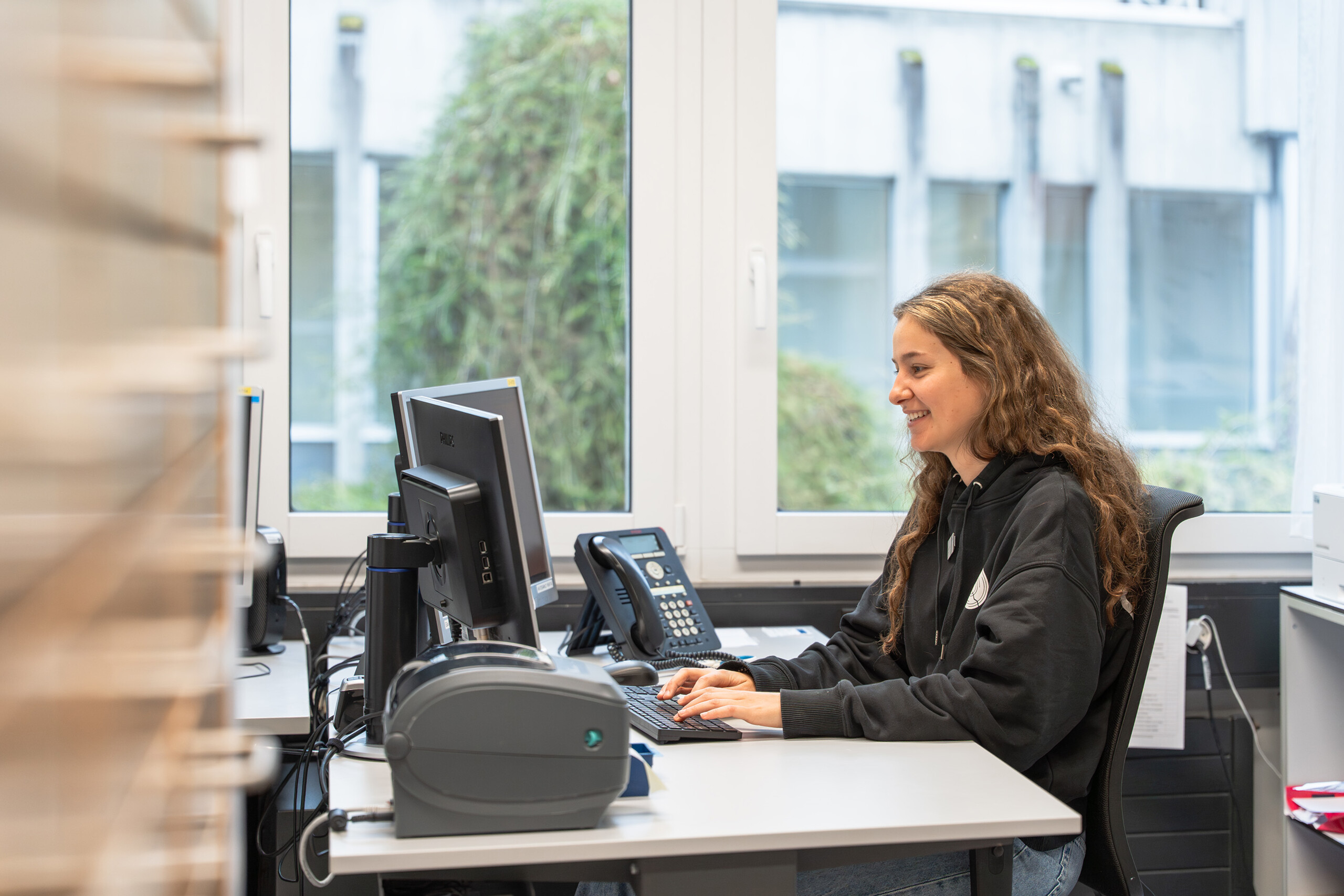 Mitarbeiterin sitzt an einem Büroarbeitsplatz mit Monitor, Telefon und Tastatur vor großen Fenstern.