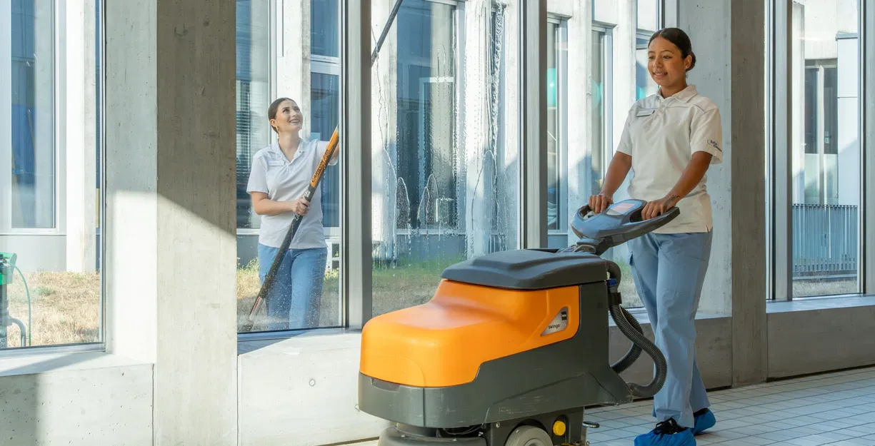 Reinigungskraft bedient eine orange-graue Scheuersaugmaschine in einem hellen Gebäude; im Hintergrund wischt eine weitere Reinigungskraft eine Glasfront.