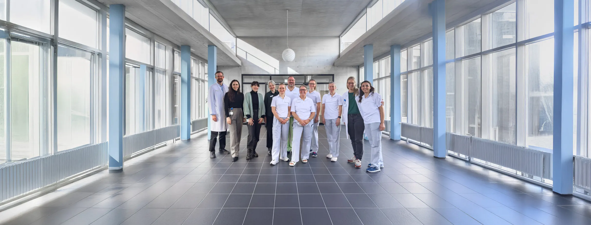 Gruppe von neun Personen in einem modernen, hellen Raum mit großen Fenstern. Die Anwesenden tragen unterschiedliche Kleidung, darunter medizinische Uniformen. Der Raum hat einen grauen Boden und blaue Wände, mit einer minimalistischen Deckenleuchte.