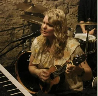 Frau mit lockigem blondem Haar spielt eine schwarze Gitarre vor einer Drumset-Bühne in einem dunklen, rockigen Setting