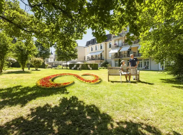 Zwei Personen sitzen auf einer Bank im Garten, umgeben von Bäumen und Rasen. Im Vordergrund ist ein farbenfrohes Blumenarrangement in Form einer geschwungenen Linie zu sehen. Im Hintergrund stehen Gebäude mit Balkonen und Sonnenschirmen.