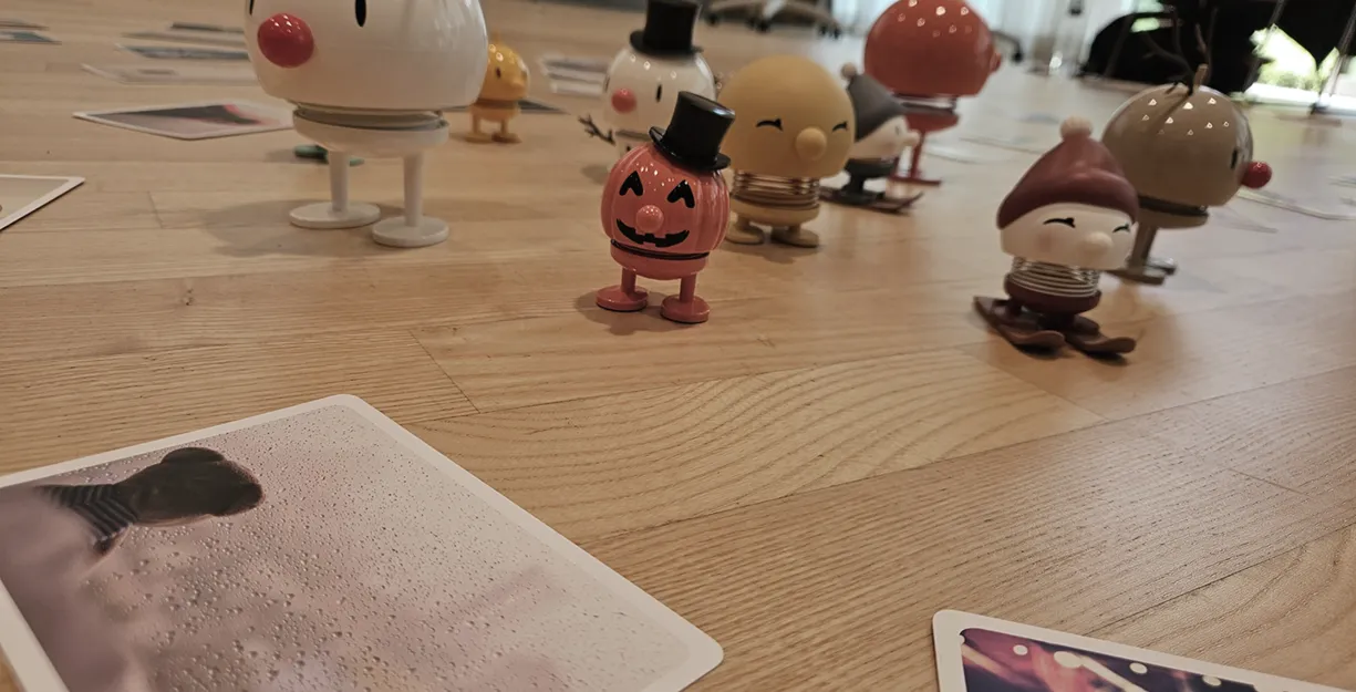 Bunte Figuren in verschiedenen Designs stehen auf einem Holzboden, umgeben von mehreren Fotos. Eine Figur hat ein Rentiergeweih, eine andere trägt einen Zylinder, und eine dritte hat einen roten Hut. Die Szene wirkt verspielt und fröhlich.