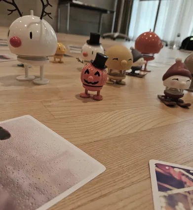 Bunte Figuren in verschiedenen Designs stehen auf einem Holzboden, umgeben von mehreren Fotos. Eine Figur hat ein Rentiergeweih, eine andere trägt einen Zylinder, und eine dritte hat einen roten Hut. Die Szene wirkt verspielt und fröhlich.