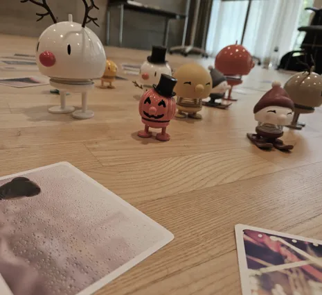 Bunte Figuren in verschiedenen Designs stehen auf einem Holzboden, umgeben von mehreren Fotos. Eine Figur hat ein Rentiergeweih, eine andere trägt einen Zylinder, und eine dritte hat einen roten Hut. Die Szene wirkt verspielt und fröhlich.