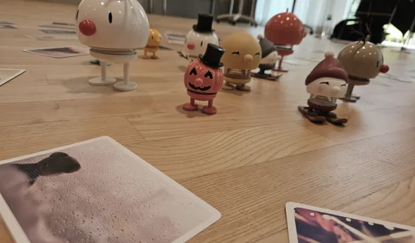 Bunte Figuren in verschiedenen Designs stehen auf einem Holzboden, umgeben von mehreren Fotos. Eine Figur hat ein Rentiergeweih, eine andere trägt einen Zylinder, und eine dritte hat einen roten Hut. Die Szene wirkt verspielt und fröhlich.