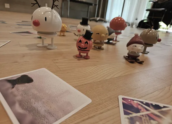 Bunte Figuren in verschiedenen Designs stehen auf einem Holzboden, umgeben von mehreren Fotos. Eine Figur hat ein Rentiergeweih, eine andere trägt einen Zylinder, und eine dritte hat einen roten Hut. Die Szene wirkt verspielt und fröhlich.