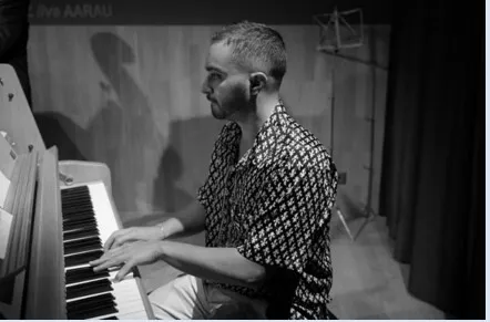 Junger Pianist an einem Flügel, von der Seite fotografiert, spielt mit den Händen auf den Tasten; Hintergrund mit Vorhang und Scheinwerfer, schwarz-weiß.