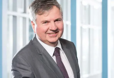 Mann in einem grauen Anzug mit Krawatte, lächelnd vor einem hellen, modernen Hintergrund mit großen Fenstern.