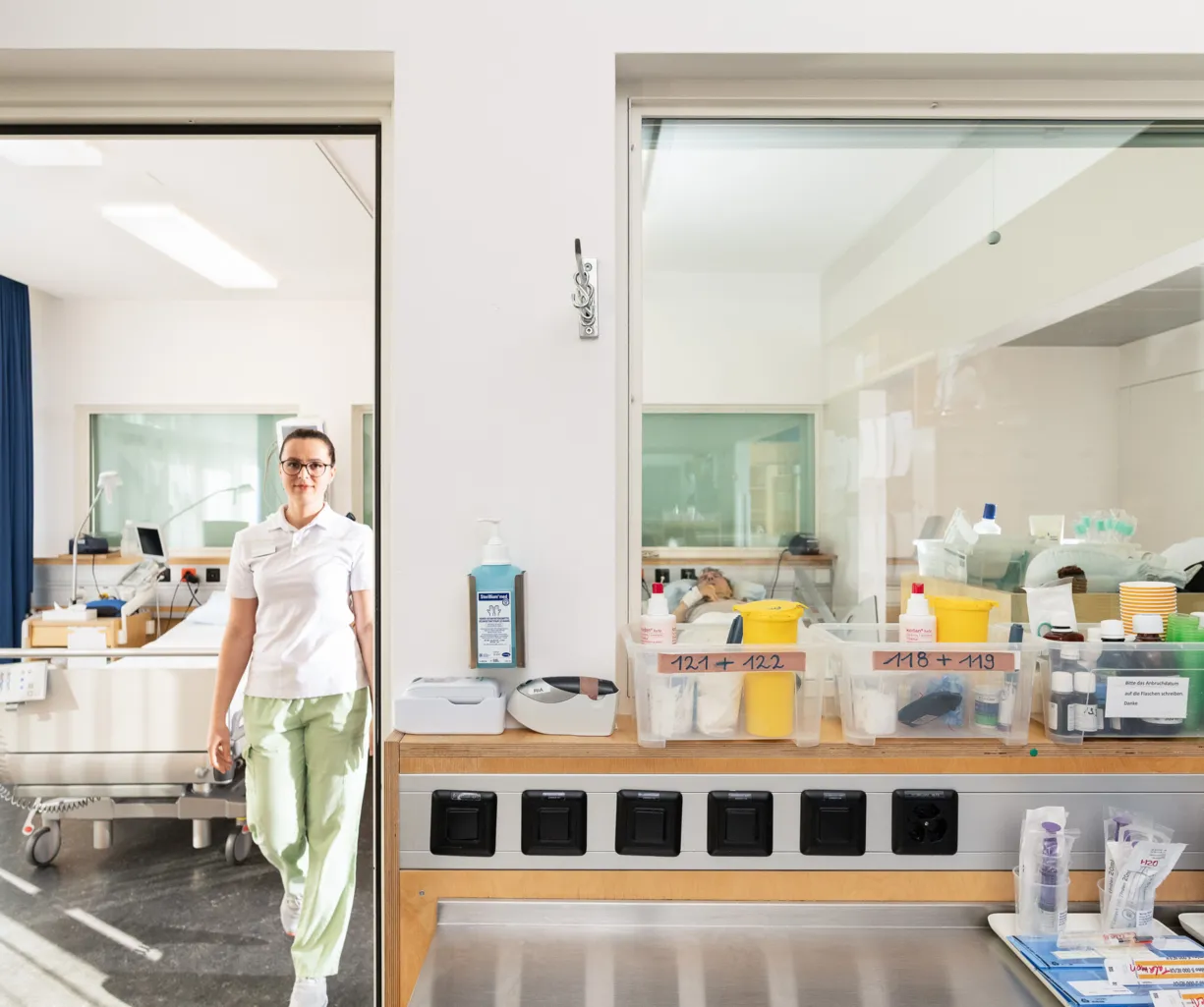 Pflegekraft in einem Krankenhaus, die in einem Türrahmen steht. Im Hintergrund sind ein Krankenhausbett und medizinische Geräte sichtbar. Auf einem Tisch sind verschiedene Behälter mit Medikamenten und Pflegeutensilien angeordnet.