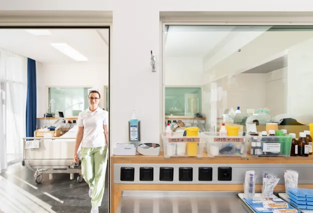 Pflegekraft in einem Krankenhaus, die in einem Türrahmen steht. Im Hintergrund sind ein Krankenhausbett und medizinische Geräte sichtbar. Auf einem Tisch sind verschiedene Behälter mit Medikamenten und Pflegeutensilien angeordnet.