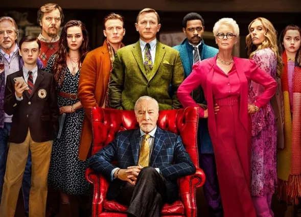 Gruppe von Personen in formeller Kleidung, die um einen älteren Mann in einem roten Sessel stehen. Der Mann trägt einen Anzug mit Krawatte und hat graue Haare. Die anderen Personen zeigen verschiedene Gesichtsausdrücke und Stile, darunter eine Frau in...