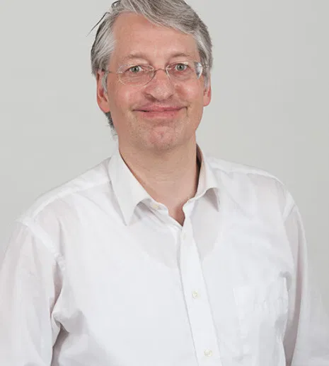 Porträt eines lächelnden Mannes mit grauem Haar und Brille in weißem Hemd vor hellem Hintergrund.