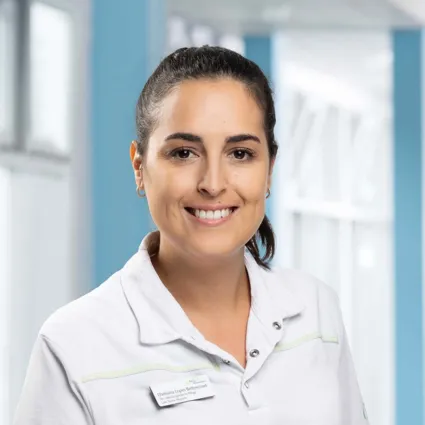 Lächelnde Frau mit langen, dunklen Haaren, trägt ein helles Oberteil mit einem Namensschild, im Hintergrund ein heller Raum mit blauen Wänden und Fenstern.