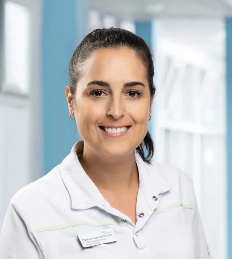 Lächelnde Frau mit langen, dunklen Haaren, trägt ein helles Oberteil mit einem Namensschild, im Hintergrund ein heller Raum mit blauen Wänden und Fenstern.