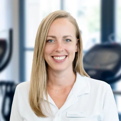 Lächelnde blonde Frau im weißen Kittel, Porträt vor unscharfem Fitnessstudio mit Trainingsgeräten, helles Hintergrundlicht.