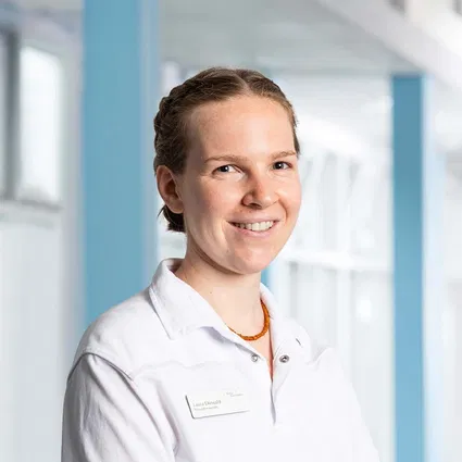 Lächelnde Frau mit zurückgebundenen Haaren im weißen Kittel und orangefarbener Halskette vor hellem, modernem Hintergrund.