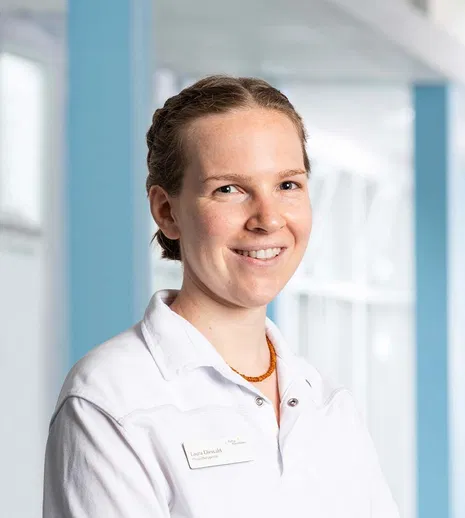 Lächelnde Frau mit zurückgebundenen Haaren im weißen Kittel und orangefarbener Halskette vor hellem, modernem Hintergrund.