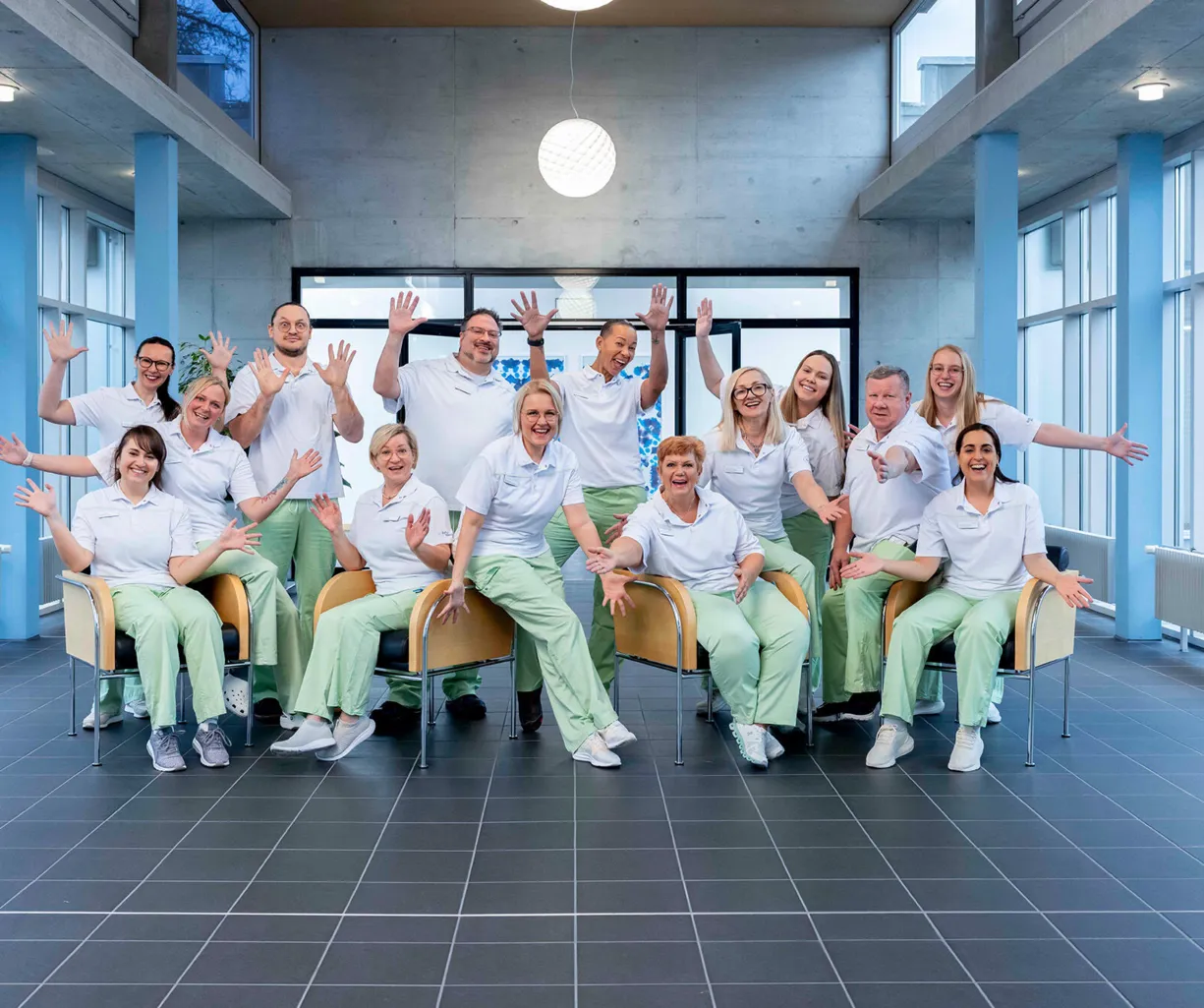 Gruppe von 14 Personen in weißen Oberteilen und grünen Hosen, die in einem modernen Raum mit großen Fenstern stehen und sitzen. Sie lächeln und heben die Hände in die Luft, um Freude auszudrücken. Der Boden ist gefliest, und eine runde Lampe hängt von...