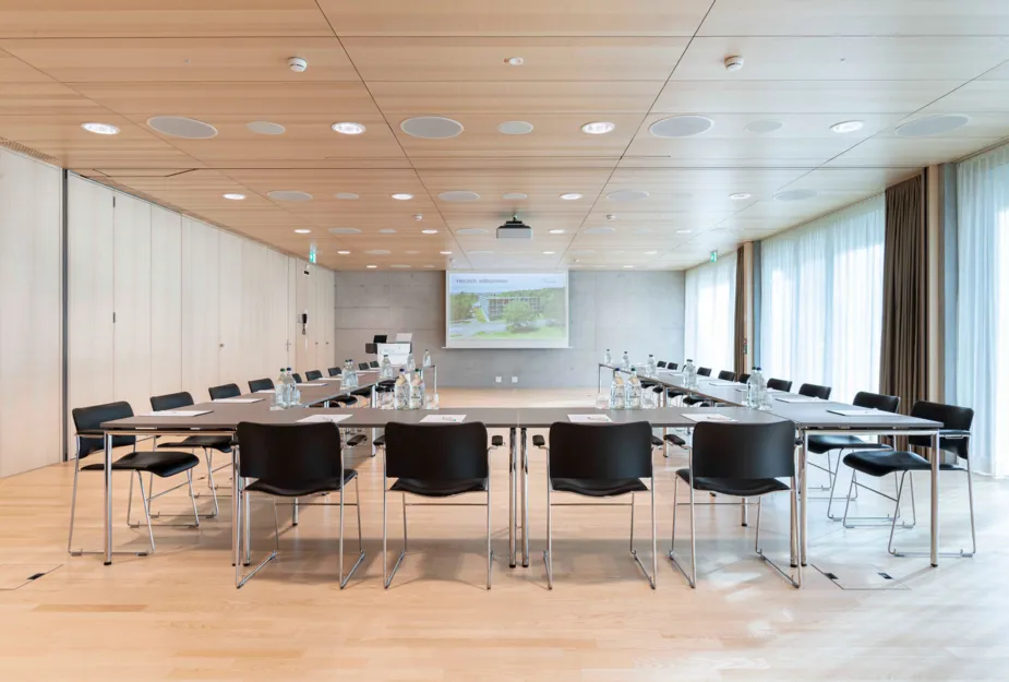 Konferenzraum mit einem langen Tisch, umgeben von schwarzen Stühlen, ausgestattet mit Wasserflaschen und Notizblättern. An der Wand ein Projektor mit einer Präsentation, große Fenster mit Vorhängen und Holzdecke.