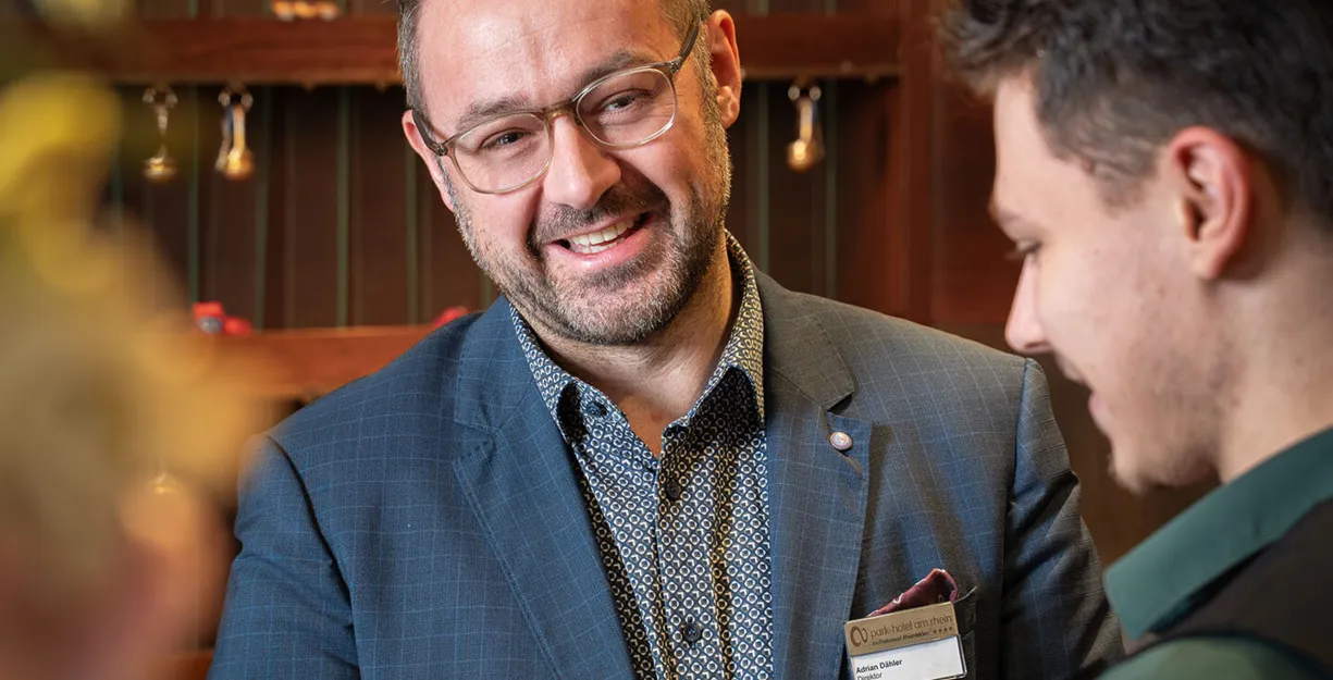 Zwei Männer schütteln sich die Hände in einem eleganten Innenraum. Der Mann links trägt einen blauen Anzug mit einem gemusterten Hemd und einer Namensschilder, während der Mann rechts ein grünes Hemd trägt. Im Hintergrund sind dekorative Elemente und e...