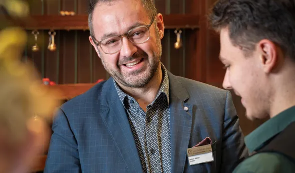 Zwei Männer schütteln sich die Hände in einem eleganten Innenraum. Der Mann links trägt einen blauen Anzug mit einem gemusterten Hemd und einer Namensschilder, während der Mann rechts ein grünes Hemd trägt. Im Hintergrund sind dekorative Elemente und e...