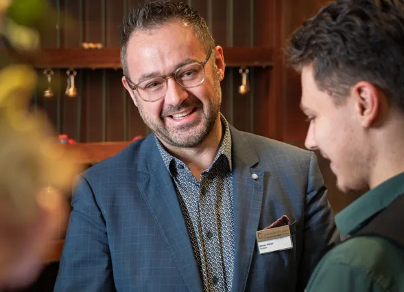 Zwei Männer schütteln sich die Hände in einem eleganten Innenraum. Der Mann links trägt einen blauen Anzug mit einem gemusterten Hemd und einer Namensschilder, während der Mann rechts ein grünes Hemd trägt. Im Hintergrund sind dekorative Elemente und e...