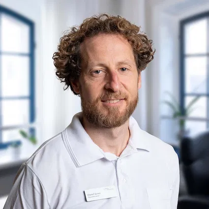 Porträt eines lächelnden Mannes mit lockigem Haar und Bart in weißem Poloshirt mit Namensschild, vor hellem Bürohintergrund.