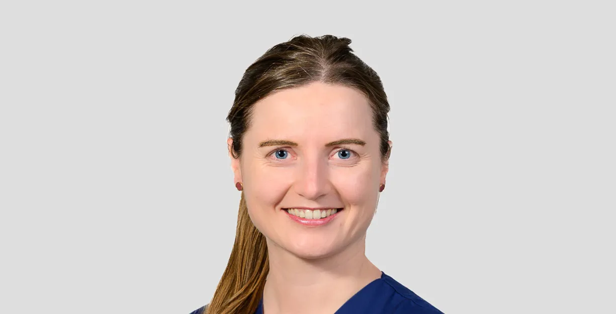 Frau mit langen, braunen Haaren in blauer Arbeitskleidung, lächelt freundlich. Ein Namensschild ist sichtbar. Hintergrund in neutralem Grau.